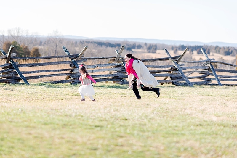 Mom playfully chasing daughter in a field by a wooden fence row.